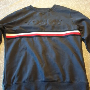 Mörkblå sweatshirt från Tommy Hilfiger  - Säljer en mörkblå sweatshirt från Tommy Hilfiger helt oanvänd  med upphöjd TOMMY-text på bröstet och en rand i vitt, rött och blått. Klassisk rund halsringning och långa ärmar. Perfekt för en stilren och avslappnad look