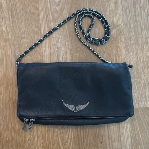 Svart axelväska Zadig Voltaire - Säljer en svart axelväska från märket Zadig Voltaire i skinn med silverfärgad kedjerem och snygg vingdetalj framtill. Väskan har dragkedja nertill och är perfekt för att bära det viktigaste. Stilren och passar till många outfits. Väskan saknar ett fäste för bandet på ena sidan men går ändå bra att använda. Kan säkerligen repareras. Även emblemet på utsidan är slitet, se bilder.