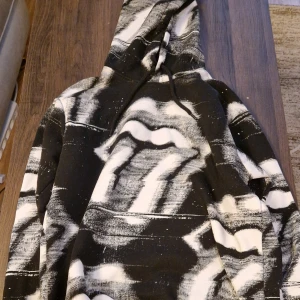 Rolling Stones hoodie - Säljer en svart hoodie med vit abstrakt print och luva. Tröjan har en stor magficka och dragsko i luvan. Passar perfekt för dig som gillar streetwear och vill sticka ut med ett unikt mönster.