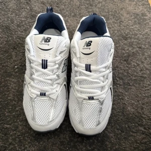 Vita New Balance sneakers - Säljer ett par fräscha vita New Balance sneakers med marinblå detaljer. Skorna har meshpaneler för extra andningsförmåga, snörning och en bekväm, platt sula. Perfekta för träning eller vardagsstil. Klassisk New Balance-logga på sidan. Helt nya.