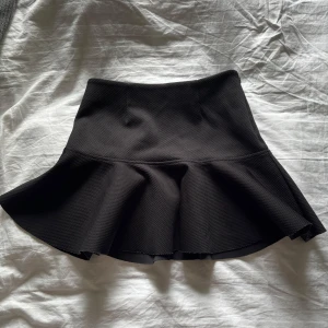 Svart kort volangkjol - En svart kortkjol med volang nertill. Kjolen har en markerad midja och är sydd i ett strukturerat tyg som ger extra stadga och form. Perfekt för dig som gillar en feminin och trendig stil.