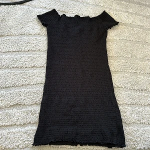 Svart smockad klänning från Gina Tricot - Svart smockad klänning från Gina Tricot med offshoulder-modell och kort ärm. Klänningen är figurnära och har en vågig kant upptill och nedtill. Perfekt för dig som gillar en enkel men trendig stil.