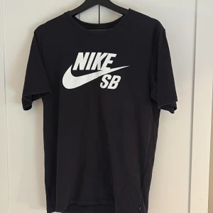 Svart Nike SB t-shirt med stor logga - Svart t-shirt från Nike SB med stor vit logga framtill. Klassisk rund hals och korta ärmar. Tillverkad i Dri-Fit material som andas och håller dig torr. Perfekt för träning. Enkel och stilren design. Hör av dig vid intresse!💕