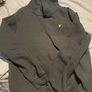 Svart hoodie från Lyle & Scott - Svart hoodie från Lyle & Scott med klassisk gul broderad logga på bröstet. Tröjan har lång ärm, huva och magficka. Perfekt för en avslappnad och stilren look.använd några gånger men ändå riktigt bra skick.hör av er vid funderingar.