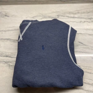 Blå hoodie från Polo Ralph Lauren - Mysig blå hoodie från Polo Ralph Lauren med vit kontrastsöm och klassisk broderad logga på bröstet. Tröjan har huva och en stor magficka framtill, perfekt för en avslappnad stil.
