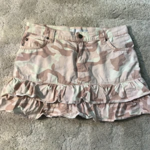 Camo minikjol med volang från Amisu - Säljer en kort camouflagemönstrad kjol från Amisu med volangkant nertill. Kjolen har klassiska jeansfickor fram och bak samt knapp och dragkedja i midjan. Perfekt för dig som gillar streetstyle och vill sticka ut lite extra.
