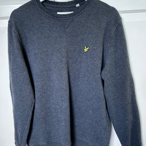 Mörkgrå sweatshirt från Lyle & Scott - Mörkgrå sweatshirt med rund hals och klassisk gul logga på bröstet från Lyle & Scott. Tröjan har långa ärmar och är tillverkad i mjuk bomullsmix. Perfekt för en avslappnad stil. Hör av er vid intresse/ frågor!