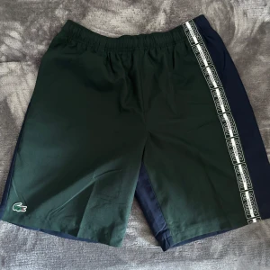 Gröna och marinblå shorts/badshorts från Lacoste - Snygga shorts från Lacoste i grönt och marinblått med vit logotypdetalj längs ena sidan och broderad krokodil på benet. Köptes på Hede fashion outlet, bara testat dom! 