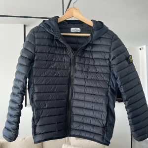 Svart dunjacka från Stone Island - Säljer en svart dunjacka från Stone Island med huva och dragkedja. Jackan har quiltad design och klassisk logotyp på ärmen. Perfekt för kyliga dagar och har en sportig look. Jackan är helt ny och aldrig använd, säljer denna eftersom den var för liten när jag fick den. Jackan är lite stor i storleken. Hör av er om ni har några frågor. Pris kan diskuteras. 