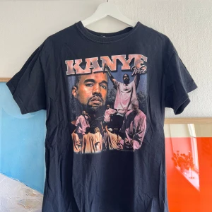 Svart Kanye West t-shirt med tryck - Cool svart t-shirt med stort Kanye West-tryck både fram och bak. Texten 'Kanye West' och 'God Show Me The Way' syns tydligt. Tillverkad i 100% bomull och har normal passform med korta ärmar.