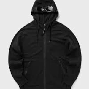 Svart hoodiejacka med goggles från C.P. Company - Säljer en svart hoodiejacka från C.P. Company med dragkedja och ikoniska goggles i huvan. Jackan har två fickor med dragkedja och justerbar huva med snörning. Perfekt för dig som gillar streetwear och vill sticka ut.