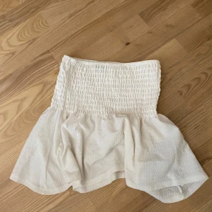 Vit kortkjol från Gina Tricot - Supersöt vit kortkjol från Gina Tricot med smockad midja och volang nertill. Kjolen är tillverkad i ett lätt och luftigt material, perfekt för varma dagar. Enkel att matcha med olika toppar för en fräsch look.