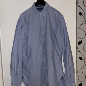 Blå oxfordskjorta från Polo Ralph Lauren - Klassisk ljusblå oxfordskjorta från Polo Ralph Lauren med button-down krage och broderad logga på bröstet. Skjortan har långa ärmar och knäppning framtill. Perfekt till både jeans och chinos.