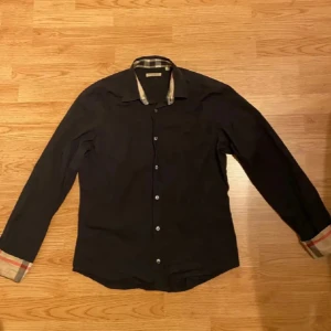 Svart skjorta från Burberry Brit med rutiga detaljer - Säljer en svart skjorta från Burberry Brit i storlek M. Skjortan har klassiska rutiga detaljer på insidan av kragen och ärmsluten, samt knäppning framtill. Perfekt för dig som gillar stilrena plagg med en twist.
