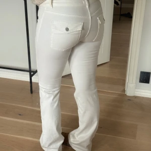 Vita bootcut jeansbyxor med fickdetaljer - Säljer ett par vita jeansbyxor med bootcut passform och  fickor bak med knappdetalj. Byxorna har låg midja och markerade sömmar. Använda en gång då det inte riktigt var i min smak, men skit snygga!!🫶🏼