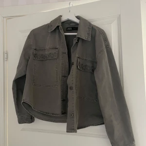 Grå/grön overshirt från Zara - Säljer en grå/grön overshirt från Zara med två stora bröstfickor och dekorativ brodyr på kragen. Jackan har knäppning framtill och är tillverkad i ett kraftigare bomullstyg. Perfekt att slänga över en hoodie eller t-shirt.