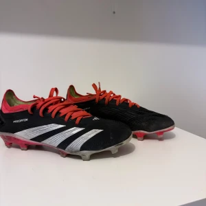 Adidas Predator fotbollsskor med röda snören - Säljer ett par Adidas Predator fotbollsskor i svart med vita detaljer och röda snören. Skorna har låg profil och är designade för spel på gräs med fasta dobbar. Perfekta för dig som vill ha bra grepp och kontroll på planen. De är använda men i bra skick. Skorna kommer såklart att rengöras innan fraktning. Pris kan diskuteras