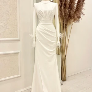 Vit långklänning med fjäderdetaljer - Elegant vit långklänning med lång ärm och hög hals. Klänningen har draperad midja och fjäderdetaljer vid ärmslut. Perfekt för speciella tillfällen där du vill sticka ut med en stilren look.