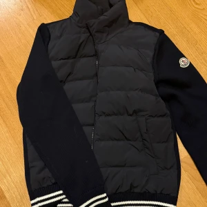 Moncler Cardigan - Snygg svart pufferjacka från Moncler med stickade ärmar och ribbade muddar med vita ränder. Jackan har dragkedja framtill och klassisk Moncler-logga på ärmen. Perfekt för kyliga dagar och stilren look. Passar S/M