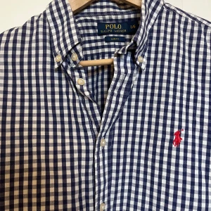 Blårutig skjorta från Polo Ralph Lauren - Klassisk blårutig skjorta från Polo Ralph Lauren med röd broderad logga på bröstet. Skjortan har lång ärm, knappar framtill och button-down krage. Slim fit-modell i mjuk bomull. Väldigt bra skick, passar som en medium!