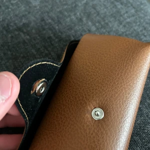 Brunt Ray-Ban glasögonfodral - Snyggt brunt glasögonfodral från Ray-Ban i läderimitation med tryckknapp och logotyp på framsidan. Perfekt för att skydda dina solglasögon eller vanliga glasögon när du är på språng. Enda problemet är när man öppnar och stänger har den lossnat, men fungerar ändå. Passar till ray ban boyfriends storlek 56.