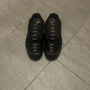 Nike Air Max Plus TN helsvarta sneakers - Säljer ett par helsvarta Nike Air Max Plus TN sneakers med klassisk TN-logga på hälen och synliga Air Max-dämpningar. Skorna har mesh och syntetpaneler, svarta detaljer och diskret Nike Swoosh på sidan. Perfekta för dig som gillar sportig och stilren look.