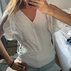 Vit blus med v-ringning och korta ärmar - Säljer en luftig vit blus med v-ringning och korta, vida ärmar. Blusen har en avslappnad passform och knytband vid axlarna för extra detalj. Perfekt till sommaren och enkel att matcha med jeans eller kjol.