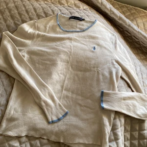 Vit långärmad tröja från Ralph Lauren - Säljer en vit långärmad tröja från Ralph Lauren med ljusblå detaljer vid halsringning och ärmslut. Tröjan har en liten broderad logga på bröstet och är tillverkad i ett tunt, mjukt material. Fint skick förutom en lagning på framsidan