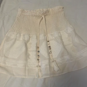 Beige kortkjol med smock och broderi - Söt beige kortkjol med smock i midjan och dekorativa broderier i vitt. Kjolen har knytband med pärldetaljer och tofsar framtill. Perfekt för en somrig och avslappnad stil.