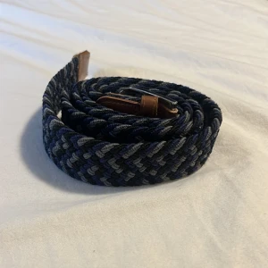 Flätat bälte - Snyggt flätat bälte i mörkblå, grå och svarta toner med bruna skinn-detaljer vid spännet och änden. Spännet är i silverfärgad metall. Jag tror att storleken är 80 kanske 70 