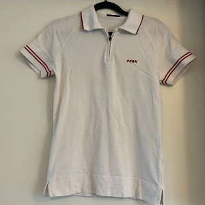 Vit pikétröja med dragkedja från Peak Performance - Vit pikétröja från Peak Performance med röda ränder på ärmar och krage. Tröjan har korta ärmar och dragkedja framtill. Klassisk piké i bomull, perfekt för en sportig stil. Storlek M är för ungdom. En liten missfärgning, syns knappt. Se bild 4.