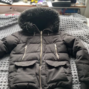 Svart pufferjacka med fuskpäls från Supply & Demand - Säljer en svart pufferjacka från Supply & Demand med stor huva och fuskpäls. Jackan har dragkedja framtill, två snedställda bröstfickor med dragkedja och två fickor med lock. Perfekt för kalla dagar!