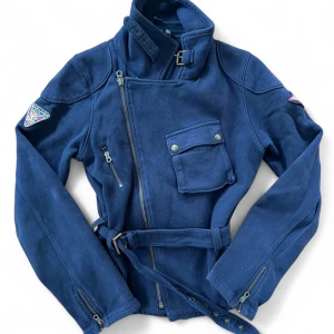 Mörkblå jacka med bälte - Säljer en mörkblå jacka med sned dragkedja, bälte i midjan och dekorativa patchar på ärmarna. Jackan har en ficka med lock och knappar på bröstet samt dragkedjor vid ärmsluten. Snygg och cool modell med hög krage.