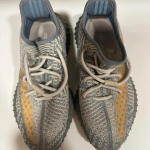 Adidas Yeezy Boost 350 V2 sneakers i grå och beige - Säljer ett par äkta Adidas Yeezy Boost 350 V2 sneakers med stickad ovandel i grått och beige med inslag av gulorange längs sidan. Skorna har rund tå, platt sula och klassisk snörning. Sulan är i gummi med boost-dämpning för extra komfort. Kartongen är dock slängd därav priset men de går att diskutera vid snabb affär och jag har bilder från när skorna var nya med kartogen!