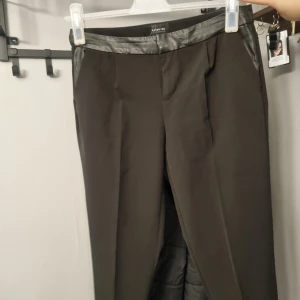 Svarta kostymbyxor med skinnliknande detaljer från Reserved - Snygga svarta kostymbyxor från Reserved med skinnliknande detaljer i midjan och vid fickorna. Byxorna har raka ben och klassisk passform, perfekta för en stilren look.