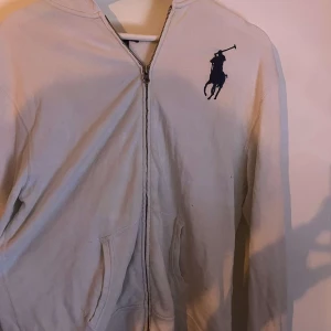 Vit hoodie med dragkedja från Polo Ralph Lauren - Vit hoodie från Polo Ralph Lauren med dragkedja framtill och två fickor. Stor blå logga på bröstet. Klassisk huva och långärmad modell, perfekt för en avslappnad stil.