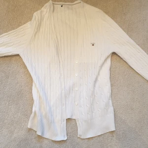 Vit kabelstickad kofta från Gant - Vit kabelstickad kofta från Gant med knappar framtill och broderad logga på bröstet. Klassisk design med ribbade muddar och lång ärm. Perfekt för lager-på-lager och enkel att matcha med olika outfits.