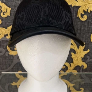 Svart keps från Gucci  - Snygg svart keps från Gucci med diskret GG-logga i ton-i-ton över hela kepsen. Klassisk böjd skärm och mesh-liknande material som ger en cool vibe. Perfekt accessoar för att lyfta din streetstyle.