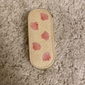 Beige liten necessär/plånbok med broderadel snäckor - Söt beige plånbok i flätad stil med broderade rosa snäckor på framsidan. Plånboken har dragkedja runtom och är perfekt för småsaker eller kort. Den har en mjuk insida och kompakt form som är lätt att ta med i väskan.
