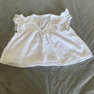 Vit volangblus från Gina Tricot - Supersöt vit blus från Gina Tricot med volangdetaljer på axlarna och smock upptill. Luftig och lätt i bomull, perfekt för varma dagar. V-ringad och ärmlös design som ger en fräsch och trendig look.