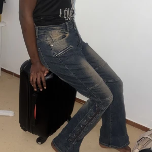 Mörka jeans med snörning och dragkedjor - Säljer ett par mörkblå jeans med coola slitningar, snörningsdetaljer längs benen och dragkedjor vid fickorna. Byxorna har hög midja och bootcut-modell som ger en snygg siluett. Perfekta för dig som vill sticka ut med din stil.