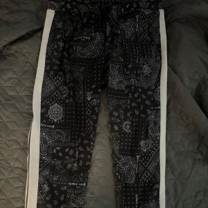 Palm angels byxor - Palm angels herr byxor med vitt bandanamönster och vita sidoränder. Byxorna har snörning i midjan och är tillverkade i ett mjukt material som passar chill eller streetwear. Snygg och unik look med tydliga detaljer och bekväm passform. Storlek M. 