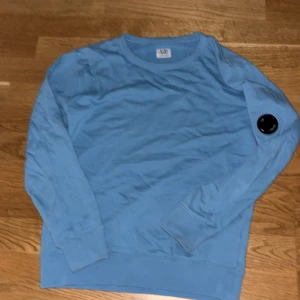 Blå crewneck från C.P. Company - Snygg blå crewneck från C.P. Company med deras ikoniska svarta lins på ärmen. Tröjan har rund hals, långa ärmar och är gjord i mjuk bomull. Perfekt för dig som gillar streetwear och vill sticka ut med en clean look.