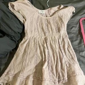 Ljusrosa lång blus  med spetsdetaljer - Supersöt ljusrosa lång blus/topp med romantiska spetsdetaljer och lätt rynkat tyg. Blusen har korta ärmar och markerad under bysten som ger en feminin siluett. Perfekt för varma dagar när du vill känna dig extra fin.