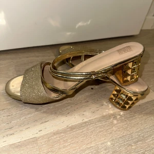 Glittriga guldiga sandaler med klack - Snygga sandaler i guld med glittrig rem över tårna och coola geometriska klackar. Öppen tå och dubbla metallicband ger en trendig vibe. Perfekta för dig som vill sticka ut med extra bling och statement-klackar.