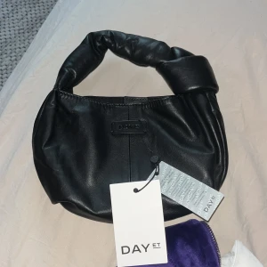 Svart handväska från DAY ET x Liva Andreasen - Helt ny!Cool svart handväska i mjukt skinn från DAY ET x Liva Andreasen. Väskan har en rundad form och ett vadderat handtag som ger en trendig look. Perfekt storlek för det viktigaste och stängs med magnetknapp. Enkel och stilren design med diskret logga framtill.