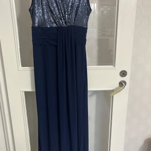 Mörkblå långklänning med paljetter  - Snygg mörkblå långklänning med glittrig paljettöverdel och draperad midja. Ärmlös modell med rund halsringning.