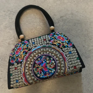 Broderad handväska med färgglada mönster - Unik handväska med färgglad brodyr i blått, rosa, gult och vitt. Väskan har svarta handtag med träpärlor och ett detaljerat cirkelmönster i mitten samt blommiga inslag. Perfekt accessoar för dig som vill sticka ut med din stil.