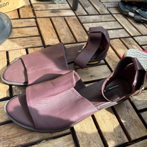 Vinröda skinnsandaler från H&M - Snygga vinröda sandaler i äkta skinn från H&M. Modellen har bred rem över foten, öppen tå och justerbart spänne vid ankeln. Platt sula och minimalistisk design som ger en clean look. Perfekta för varma dagar när du vill vara både bekväm och stilren.