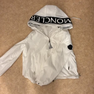 Moncler vindjacka (Pris kan diskuteras) - Snygg vit vindjacka från Moncler med stor luva och svart logga på ärmen. Jackan har dragkedja framtill, två fickor med dragkedja och coolt Moncler-tryck på huvkanten. Perfekt för dig som vill ha en clean och sportig look.